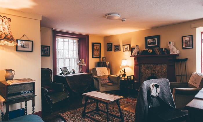 The Parlour. 1950s decorated, small room with comfy chairs and pictures on the walls. A chair with a jacket hanging over the back is positioned in fro